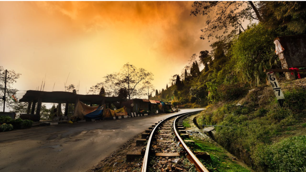 A scenic railway track winds through lush green hills, with a small village nestled along the road. The sky is ablaze with vibrant orange hues, casting a warm glow over the landscape.